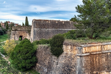 Fototapeta premium Imposing stone walls of Montjuic Castle in Girona, Spain, with angular bastions and weathered ramparts partly covered by Mediterranean vegetation and cypress trees under a clear blue sky
