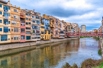 Scenic view of Girona with the iconic Eiffel Bridge over the Onyar River, Spain. Colorful riverside buildings reflect in the calm water, showcasing historic European charm