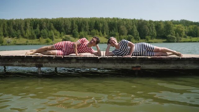 Two men in vintage striped swimwear enjoy leisure time, lying on a wooden dock by a tranquil lake, surrounded by a picturesque natural setting