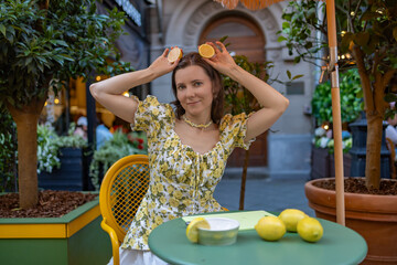 A woman in a floral dress sits at a green table, holding lemon halves on her head, surrounded by...