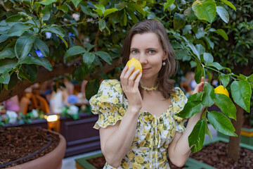 A woman in a floral dress stands under a lemon tree, holding a ripe lemon, surrounded by lush greenery