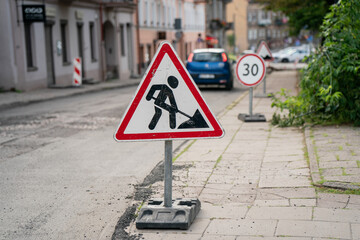 Road construction warning sign on sidewalk in residential city street with traffic limit sign and parked cars during daytime infrastructure maintenance work