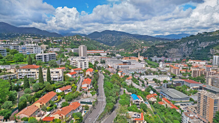 Aerial view of a vibrant urban landscape featuring residential buildings, lush greenery, winding roads, and distant mountains under a dramatic sky with clouds, showcasing city life and nature