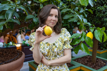 A woman in a white floral dress stands outdoors, holding a ripe lemon, surrounded by lush green trees