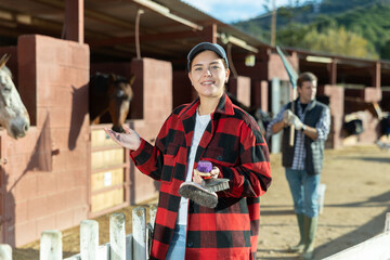 Cheerful young female in plaid shirt holding brushes for cleaning grooming horses and smiling at...