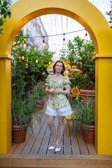 A woman in a floral dress stands under a yellow arch surrounded by lush greenery and hanging lights, holding a ripe lemon
