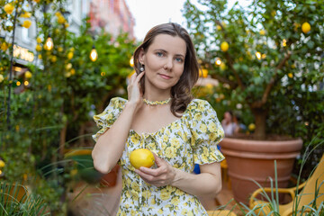 A woman in a white floral dress holds a ripe lemon in a lush garden surrounded by trees and warm lighting