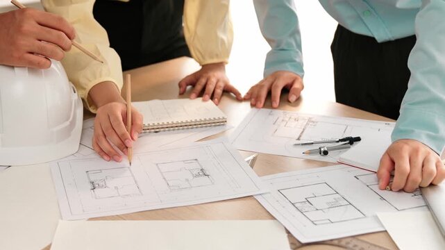 Smart architect and safety engineer discussion about building blueprint on table with architectural document, measurement tools and wooden block scatter around at modern office. Closeup. Delineation.