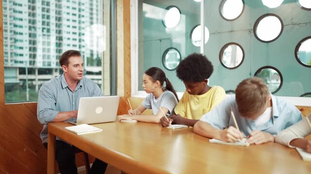 Smart teacher answer question while diverse student sitting and doing test. Multicultural children writing or taking notes while teacher using laptop analyze data and looking at paper. Edification - Powered by Adobe