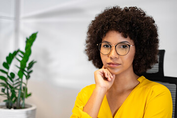 Pensive young Black woman in glasses and yellow blouse sitting at desk in home office. Thoughtful...