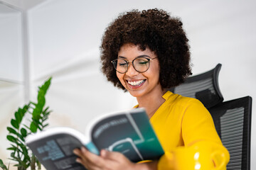Smiling Black woman reading a book at her desk in a modern office. Relaxed and inspired moment of...