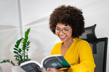 Smiling Black woman reading a book at her desk in a modern office. Relaxed and inspired moment of...
