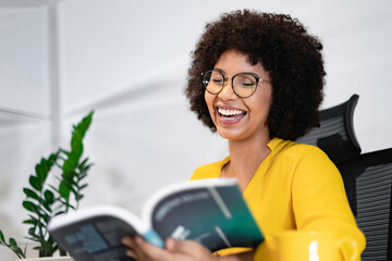 Smiling Black woman reading a book at her desk in a modern office. Relaxed and inspired moment of...