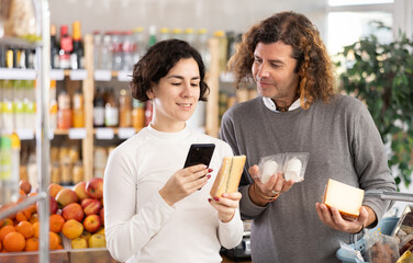 Wife scans the barcode of the cheese package on her mobile and helps her husband with the choice. Couple pays for food and purchases using a QR code in a supermarket