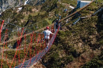 A young man wearing a helmet walks across a suspension bridge in the mountains between two peaks,...