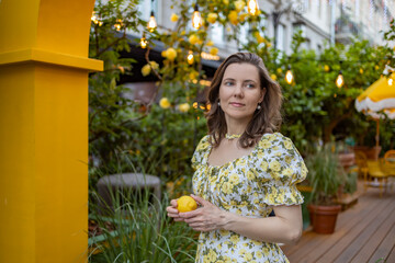 A woman in a white dress with yellow flowers holds a lemon while standing outdoors among lemon...