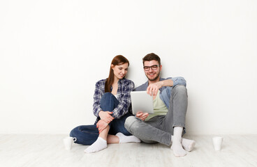 Couple shopping online on tablet, discussing purchase sitting at home floor in empty living room