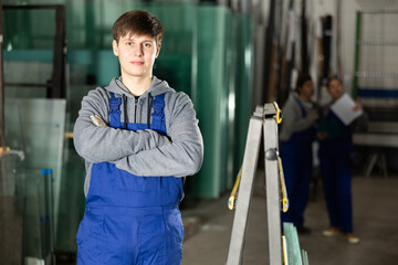 In glass workshop, young man glazer works with glass sheets and related supporting parts, preparing...