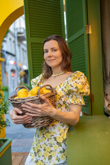 A woman in a yellow floral dress holds a basket of fresh lemons, standing in front of green shutters and an outdoor setting