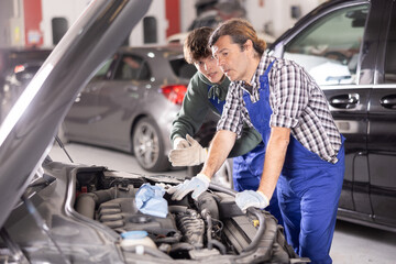 Experienced middle-aged automotive mechanic mentoring young apprentice. Men conversing while...