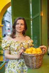 A woman in a white floral dress stands outdoors, holding a basket filled with fresh lemons, exuding a cheerful and vibrant atmosphere