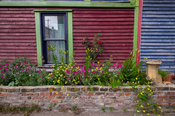 A red wooden exterior wall with a double-hung glass window. The trim around the window is painted black and green. The adjoined house is bright blue clapboard siding with a brick planter box. 