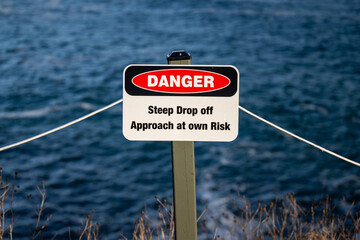 A white metal sign with danger in bold letters. There's a red and black symbol with a steep...