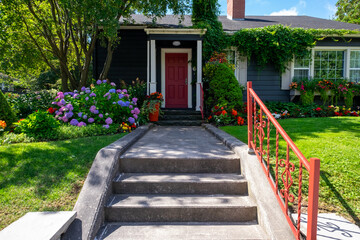A red door on a wooden navy blue house with white trim. The cottage has lush greenery around the entrance: red, yellow, pink, and white colored flowers, hydrangea shrubs, and green grass in front.
