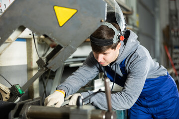 Young guy worker in safety helmet puts steel part on machine in factory