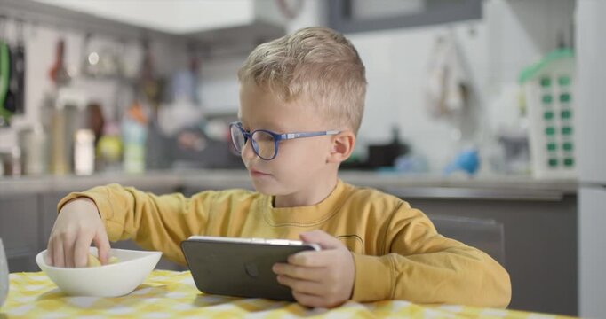 A Young Boy in Blue Glasses is Fully Engaged with his Smartphone, Taking a Break to Eat Sliced Apple Pieces While Sitting at the Kitchen Table, Illustrating Modern Childhood Habits and Screen Time.
