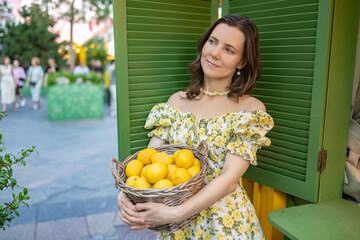 A woman in a summer floral dress with open shoulders stands outdoors, holding a basket filled with fresh lemons, against a backdrop of green shutters