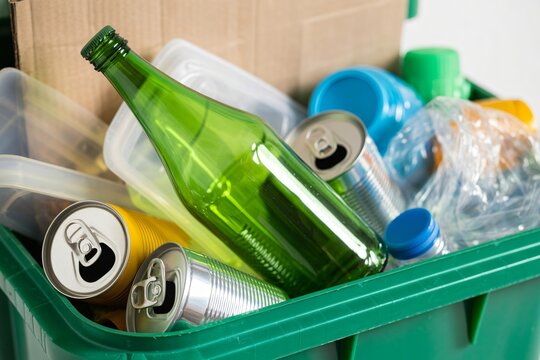 A green recycling bin is overflowing with a diverse collection of sorted recyclable materials. Clear plastic bottles, a green glass bottle, aluminum cans, and cardboard are clearly visible