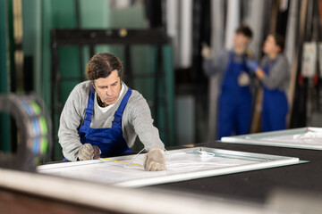 Adult man worker in uniform measuring glass sheet with tape measure on table in glass factory