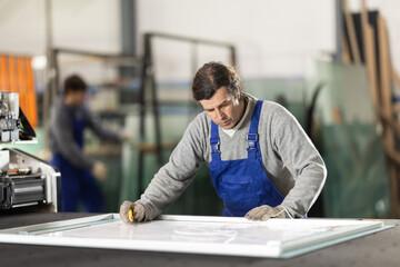 Adult man worker in uniform cutting glass sheet on table in glass factory