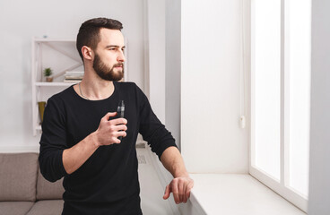Confident young bearded man vaping electronic cigarette standing against window on white...