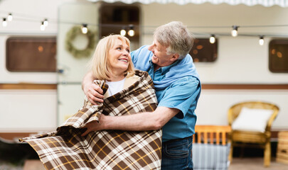 Happy senior man putting warm plaid on his beloved wife's shoulders at camping site. Panorama