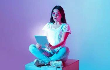  Shock at information received. Young asian woman in glasses sits in lotus position and holds tablet with luminous screen in neon © Prostock-studio