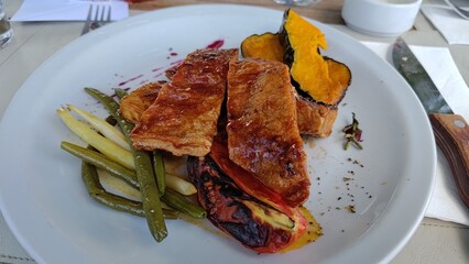 Gourmet dish with crispy pork belly, roasted pumpkin, green beans, and charred pepper on a white plate. Healthy modern cuisine served in a fancy restaurant for lunch or dinner.