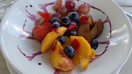 Close-up of a delicious and healthy fruit salad in a white bowl, featuring strawberries, blueberries, peaches, and grapes, garnished with edible flower petals and a sweet berry sauce.