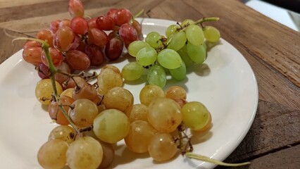 A close-up of a white plate filled with a delicious assortment of fresh red, green, and yellow grapes, showcasing a healthy and natural fruit snack on a rustic wooden table background.