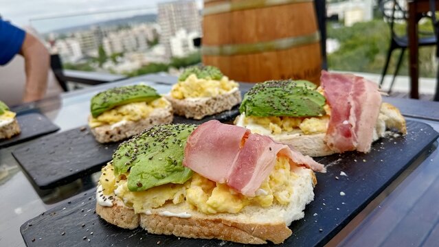 Gourmet avocado toast with scrambled eggs, bacon, and chia seeds served on a black slate platter for brunch at a rooftop cafe with a city view in the background. Healthy eating lifestyle.