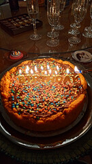 Close-up of a festive birthday cake with lit candles spelling 'BIRTHDAY', covered in chocolate icing and colorful star sprinkles. A celebration scene with champagne flutes in the background.