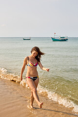 A joyful woman in a colorful bikini walks barefoot along the shoreline, with gentle waves splashing around her feet and traditional fishing boats in the background.