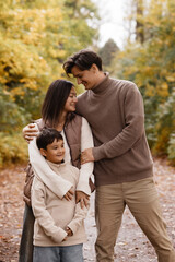 Fototapeta premium Front view of a smiling young Asian family mother, father, and son standing close in an autumn park. Casual warm clothes. Look at each other. Perfect for family, love, parenthood concepts.