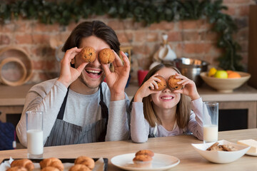 Funny young father and his daughter fooling while having snacks at kitchen, making cupcake eyes, eating homemade pastry and drinking milk
