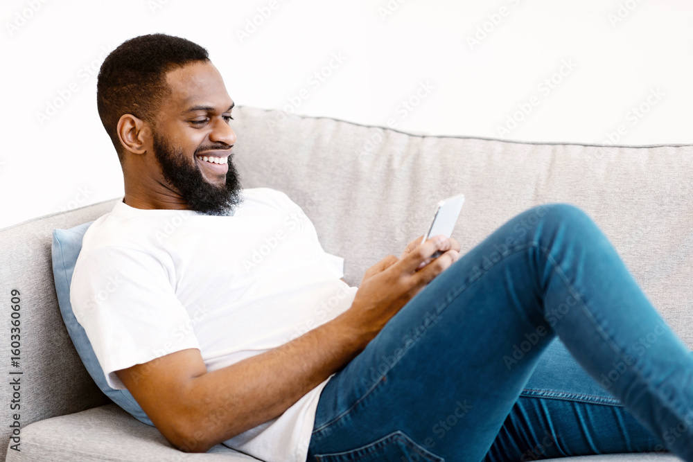 Poster mobile communication. black guy using smartphone browsing internet and texting lying on couch at hom