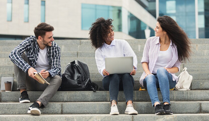 Happy students friends sitting on stairs using laptop, relaxing on urban background