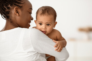 Nursery And Maternity Concept. Back view of happy African American woman with afro hair holding and hugging her small kig, enjoying spending time with lovely baby, singing lullaby to sleep