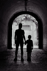 Monochrome silhouette of an adult and child holding hands at the end of a tunnel, with light behind...
