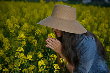 Young woman in a straw hat standing in a blooming rapeseed field, smelling a yellow rapeseed...